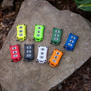 May include: A collection of eight rectangular key fob covers in various colours including neon yellow, lime green, dark green, blue, red, black, white, and orange. Each cover features three buttons with lock, unlock, and boot icons.