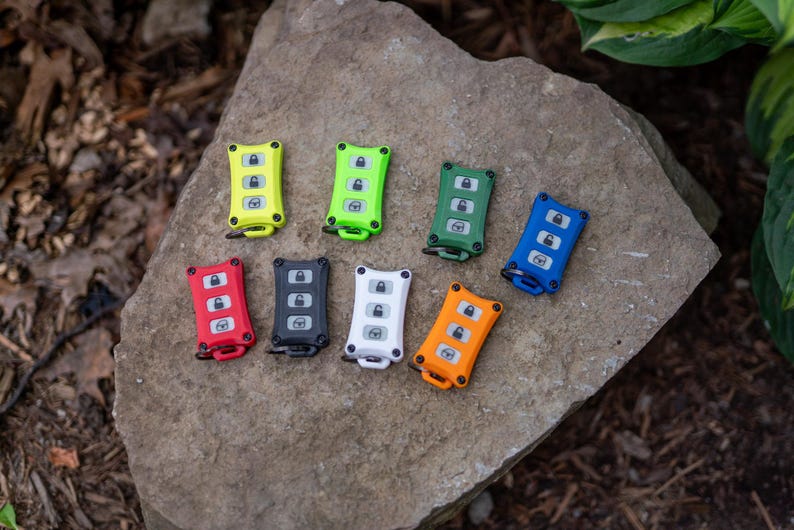 May include: A collection of eight rectangular key fob covers in various colours including neon yellow, lime green, dark green, blue, red, black, white, and orange. Each cover features three buttons with lock, unlock, and boot icons.