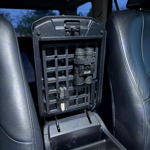 May include: Black car console organiser with a grid system. The organiser holds a pair of black binoculars labelled "10x25" and a multi-tool. The console is open, revealing the interior and the surrounding black leather seats.
