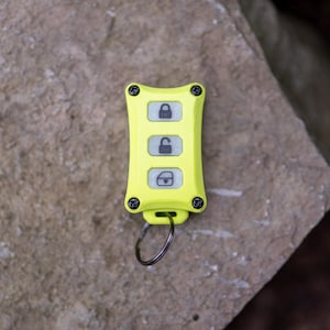 May include: A bright neon yellow rectangular key fob featuring four black corner screws and three vertical buttons. The top button displays a locked padlock icon, the middle button shows an unlocked padlock icon, and the bottom button depicts a boot icon.