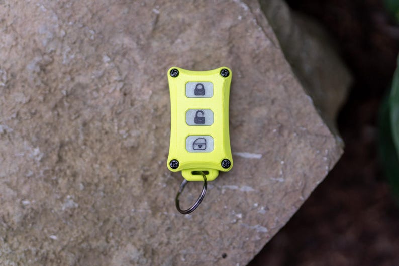 May include: A bright neon yellow rectangular key fob featuring four black corner screws and three vertical buttons. The top button displays a locked padlock icon, the middle button shows an unlocked padlock icon, and the bottom button depicts a boot icon.
