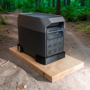 May include: A dark gray and black portable power station with multiple outlets and a digital display. It is mounted on a light brown wooden board with black supports. The background is a blurred forest path.