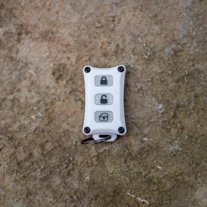 May include: A white rectangular key fob cover with four black screws at the corners. The front features three vertical buttons with black icons: a locked padlock, an unlocked padlock, and an open trunk symbol. A small metal ring is attached at the base.