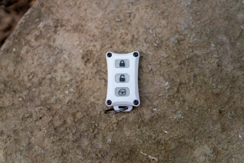 May include: A white rectangular key fob cover with four black screws at the corners. The front features three vertical buttons with black icons: a locked padlock, an unlocked padlock, and an open trunk symbol. A small metal ring is attached at the base.