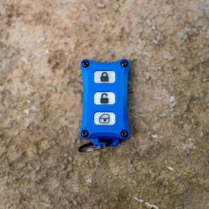 May include: A bright blue rectangular key fob cover with four black screws at the corners. The centre features three white buttons with black icons representing a locked padlock, an unlocked padlock, and a boot release. A small metal ring is attached.