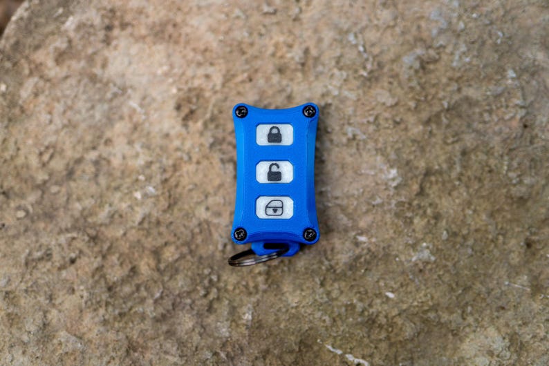 May include: A bright blue rectangular key fob cover with four black screws at the corners. The centre features three white buttons with black icons representing a locked padlock, an unlocked padlock, and a boot release. A small metal ring is attached.