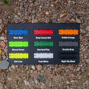 May include: A black rectangular display board featuring nine rectangular tags in various colours, each embossed with the text KodiakForged. Colours include River Blue, Deep Canyon Red, Kodiak Orange, Rescue Green, Overland Pine, Granite Gray, Volt Lime, Peak White, and Night Sky Black.