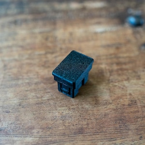 May include: A small, black rectangular plastic component with a textured surface. The component has a recessed area on one side and is likely a part of a larger device or system. The object is set against a wooden background.