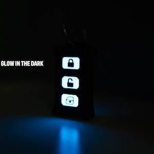 May include: A black rectangular keyring with a metal ring, featuring three white glowing buttons with icons for locking, unlocking, and a boot release. The text GLOW IN THE DARK is printed in white to the left of the keyring in a dark setting.