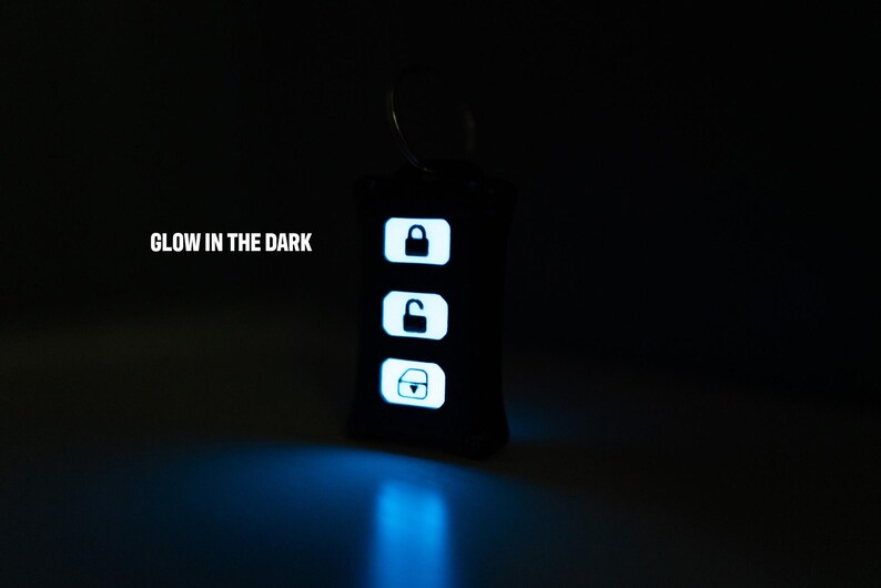 May include: A black rectangular keyring with a metal ring, featuring three white glowing buttons with icons for locking, unlocking, and a boot release. The text GLOW IN THE DARK is printed in white to the left of the keyring in a dark setting.