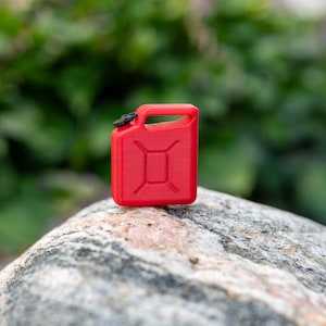 May include: A small, red miniature gas can with a black cap and handle sits on a gray rock. The gas can has a rectangular shape with a handle on top and a design on the front. The background is blurred green foliage.