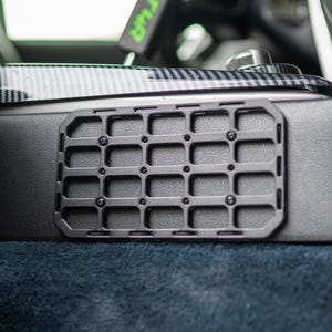 Puede incluir: Un panel MOLLE rectangular negro con un patrón de rejilla, montado sobre una superficie oscura. El panel está asegurado con pequeños tornillos negros. El fondo incluye un interior de coche con molduras de fibra de carbono y alfombra azul.
