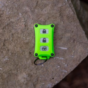 May include: A bright lime green rectangular key fob cover featuring four black screws at the corners and three white buttons with black icons representing a locked padlock, an unlocked padlock, and a boot release, attached to a black metal ring.
