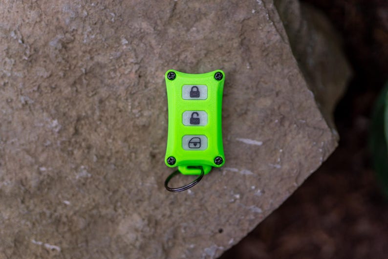 May include: A bright lime green rectangular key fob cover featuring four black screws at the corners and three white buttons with black icons representing a locked padlock, an unlocked padlock, and a boot release, attached to a black metal ring.