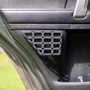 May include: A black plastic storage panel with a grid design, fitted to a car door. The panel is fixed with screws and is intended for storing small items. The door is black with a textured finish.