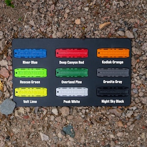May include: A black rectangular display board featuring nine rectangular tags in various colors, each embossed with the text KodiakForged. Colors include River Blue, Deep Canyon Red, Kodiak Orange, Rescue Green, Overland Pine, Granite Gray, Volt Lime, Peak White, and Night Sky Black.