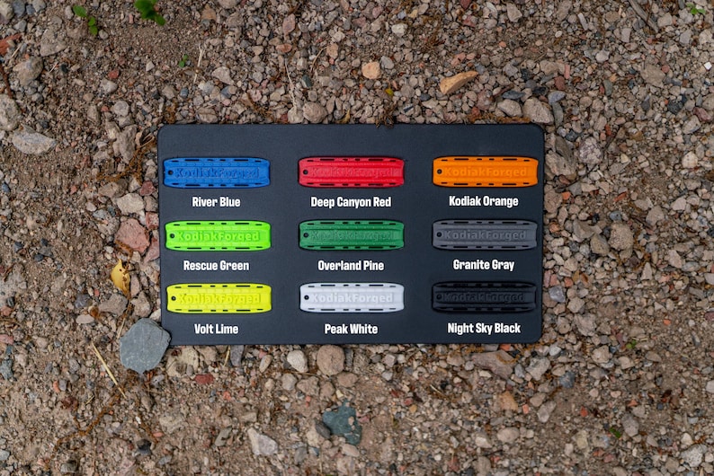May include: A black rectangular display board featuring nine rectangular tags in various colors, each embossed with the text KodiakForged. Colors include River Blue, Deep Canyon Red, Kodiak Orange, Rescue Green, Overland Pine, Granite Gray, Volt Lime, Peak White, and Night Sky Black.