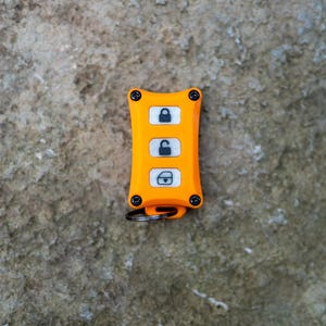 May include: A bright orange rectangular key fob cover with four black corner screws. The device features three white vertical buttons, each displaying a black icon: a locked padlock, an unlocked padlock, and a car door with a lock symbol.