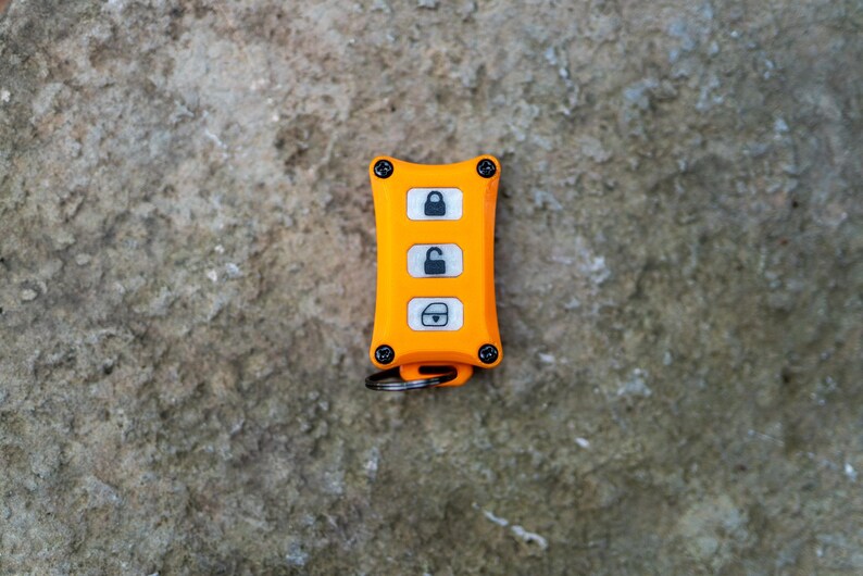 May include: A bright orange rectangular key fob cover with four black corner screws. The device features three white vertical buttons, each displaying a black icon: a locked padlock, an unlocked padlock, and a car door with a lock symbol.