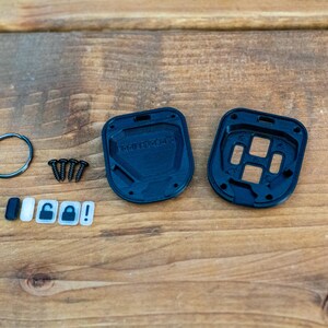May include: A black plastic key fob housing kit consisting of two interlocking shell pieces, four small metal screws, a circular metal split ring, and five small rectangular button inserts featuring icons for locking, unlocking, and an exclamation mark.