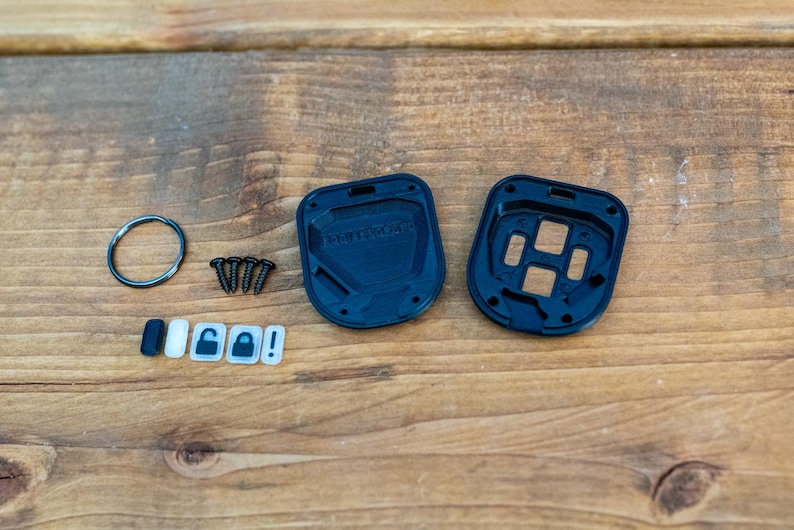 May include: A black plastic key fob housing kit consisting of two interlocking shell pieces, four small metal screws, a circular metal split ring, and five small rectangular button inserts featuring icons for locking, unlocking, and an exclamation mark.