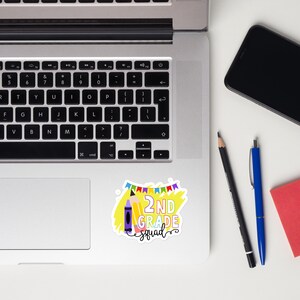 May include: A vibrant sticker featuring the words "2ND GRADE squad" in a whimsical style. The design incorporates a yellow crayon, a pencil, and a string of colourful flags. The sticker is positioned on a laptop.