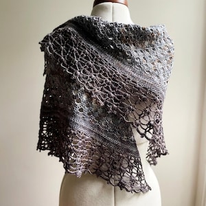 May include: A triangular crochet shawl featuring a gradient color transition from light grey to dark brown. The accessory displays an intricate openwork pattern with textured clusters and a scalloped edge, draped over a neutral-colored mannequin.