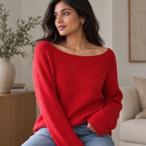 May include: A vibrant red long-sleeved sweater featuring a wide, horizontal boat neckline and a soft, knitted texture. The garment is paired with blue denim jeans, creating a simple and classic look with long sleeves and a relaxed fit.