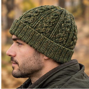 May include: A forest green knit beanie featuring a textured cable knit pattern and a ribbed cuff. The yarn is a blend of dark green and golden brown fibres, creating a variegated appearance throughout the hat. The beanie has a snug fit.