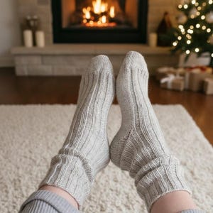 Pode incluir: Um close-up de meias cinza claro de malha canelada. As meias são usadas em ambientes internos, com uma lareira e uma árvore de Natal ao fundo. As meias são grossas e parecem quentes, perfeitas para uma noite relaxante.