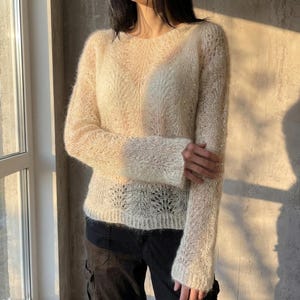 Puede incluir: Un suéter de color crema de manga larga que presenta un delicado patrón de encaje de punto abierto con un acabado texturizado y difuso. La prenda tiene un cuello redondo y una apariencia semitransparente, mostrando una estructura ligera y aireada.