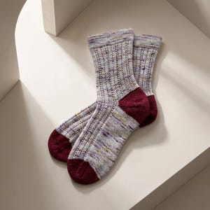 May include: A pair of mid-calf knit socks featuring a variegated light gray yarn with flecks of purple, yellow, and blue. The socks have a vertical ribbed texture throughout the leg and foot, with solid deep burgundy heels and rounded toes.