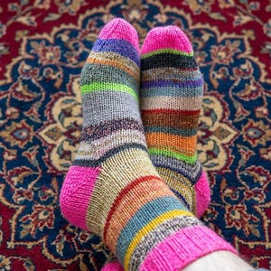 Op de afbeelding: Een paar kleurrijke, gestreepte sokken met roze hielen en manchetten. De sokken hebben banden in verschillende kleuren, waaronder roze, groen, oranje en blauw. De sokken liggen op een patroon rood en blauw tapijt.