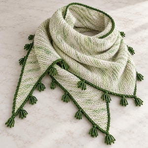 May include: A triangular knit shawl featuring a variegated pattern of cream and light green yarn. The edges are finished with a solid dark green border and decorated with small, evenly spaced dark green tassels along the perimeter.