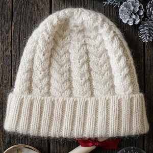 May include: Cream-colored knit beanie with a cable knit design. The hat has a ribbed brim and a textured pattern on the crown. The hat is displayed on a rustic wooden surface, with decorative elements in the background.