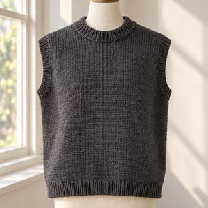 May include: A dark charcoal gray sleeveless knit sweater vest featuring a ribbed crew neckline, ribbed armholes, and a ribbed hem. The garment is displayed on a mannequin, showcasing a simple, solid-colored, textured knit construction.