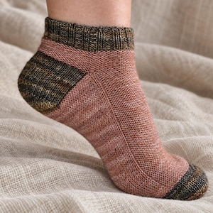 May include: An ankle sock featuring a knit construction with a dusty rose main body and dark olive green variegated yarn at the ribbed cuff, the heel, and the toe. The sock has a visible seam running along the top of the foot.