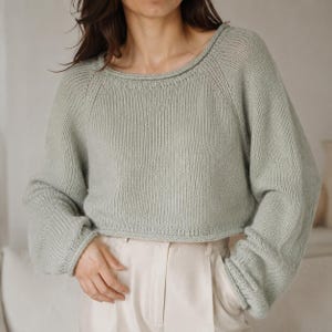 May include: A sage green long-sleeve knit jumper featuring a wide round neckline, raglan sleeves, and ribbed cuffs. The garment has a cropped length and a textured stockinette stitch pattern throughout, paired with light beige high-waisted trousers.