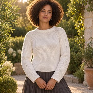 May include: A cream-colored long-sleeve knit sweater featuring a round neckline and raglan sleeves with a subtle cable knit pattern along the outer arms, paired with a dark gray skirt that reaches below the waistline in a simple design.