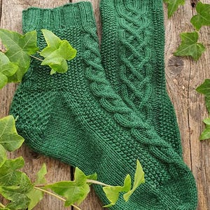 May include: A pair of emerald green knitted socks with a cable knit design on a wooden surface. The socks have a textured appearance and are adorned with green ivy leaves.