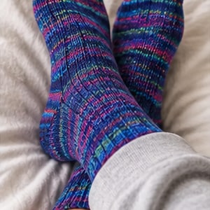 May include: A pair of hand-knitted socks featuring a vibrant variegated yarn pattern in shades of deep blue, magenta, purple, and bright yellow. The socks have a ribbed texture throughout the leg and foot, showcasing a complex, multi-colored knit design.