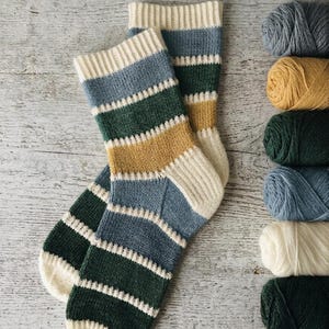 May include: A pair of knitted socks featuring horizontal stripes in cream, dusty blue, forest green, and mustard yellow. The socks have a ribbed cuff and a reinforced heel and toe section. Beside the socks are six balls of yarn in matching colours.
