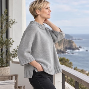 Könnte beinhalten: Ein hellgrauer Strickpullover mit geripptem Rundhalsausschnitt, Dreiviertelärmeln mit gerippten Bündchen und einem breiten gerippten Saum. Der Pullover hat eine lockere, kastige Passform mit asymmetrischem Saum und Seitenschlitzen, kombiniert mit einer dunkelgrauen Jeans.