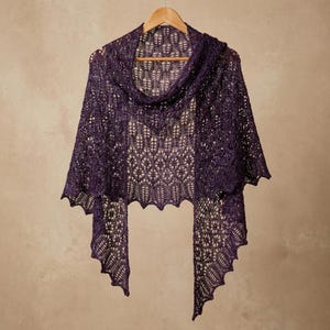 May include: A deep purple triangular shawl featuring an intricate openwork lace pattern with a scalloped edge. The garment is draped over a wooden hanger, displaying a delicate, airy knit construction with repeating leaf-like motifs throughout.