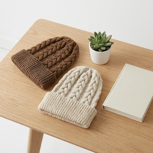 May include: Two knit beanies featuring a cable knit pattern and a ribbed cuff. One beanie is brown and the other is cream. A small green succulent in a white pot and a closed cream-colored book are positioned next to the two knit hats.