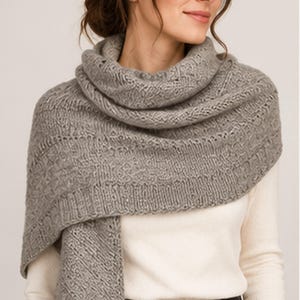 Puede incluir: Un chal de punto gris claro con un patrón texturizado, detalles intrincados tipo encaje y un borde acanalado. La prenda está drapeada alrededor del cuello y los hombros, mostrando una apariencia suave y lanosa con un color jaspeado uniforme.