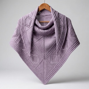 May include: A light purple triangular shawl featuring a complex knit pattern with geometric diamond shapes, eyelet details, and ribbed borders. The garment is displayed on a wooden hanger, showcasing its symmetrical construction and texture.