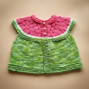 Pode incluir: Um top sem mangas tricotado à mão com um jugo rosa de padrão texturizado, separado por uma fina faixa branca de um corpo verde matizado. A peça possui uma carcela frontal com três botões esféricos brancos na linha do decote.