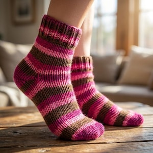 Puede incluir: Calcetines de punto con rayas rosas, marrones y magenta. Los calcetines tienen un puño acanalado y un patrón de punto texturizado. Los calcetines están sobre una superficie de madera. Los calcetines parecen hechos a mano.