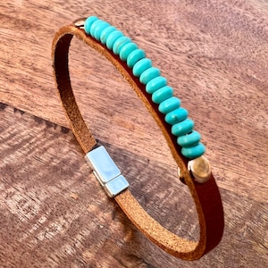 May include: A brown leather bracelet with a row of turquoise beads. The bracelet has a magnetic clasp and gold-colored rivets. The leather is a light brown color, and the beads are a bright turquoise.